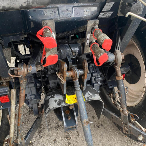 Rear view of a farming implement showing hydraulic hoses, red-handled coupling levers, and rusted metal framework on a gravel surface with a yellow warning sticker nearby
