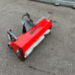Red street-sweeper attachment with white bristle roller on a paved surface, showing metal mounting brackets.