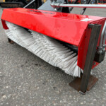 Close-up of a red industrial street broom attachment with white bristles mounted on a metal frame, resting on an asphalt surface.