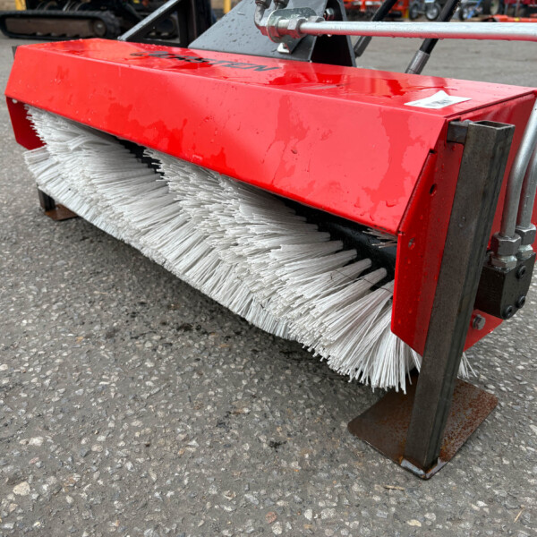 Close-up of a red industrial street broom attachment with white bristles mounted on a metal frame, resting on an asphalt surface.