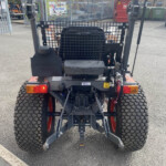 Rear view of a compact utility loader with a metal mesh cage, large off-road tires, and a hitch, parked in a lot with fences and assorted equipment in the background.