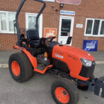 Orange Kubota tractor with roll bar parked outside a brick building near reception signs