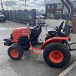 Orange Kubota tractor parked in a lot with a tall black gate and brick building in the background