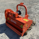 Orange Kubota agricultural implement resting on a wooden pallet in a gravel lot, with warning labels visible on the body.