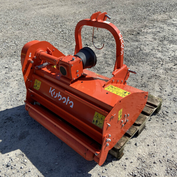 Orange Kubota agricultural implement resting on a wooden pallet in a gravel lot, with warning labels visible on the body.
