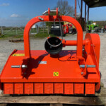 Close-up of a bright orange industrial machine frame with a large circular pulley, outdoors in a yard with other equipment.