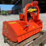 Red industrial attachment with hydraulic components, sitting on a wooden pallet in a yard; warehouse and tractors in the background.