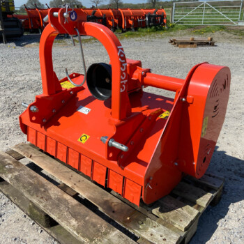 Close-up of an orange agricultural implement on a wooden pallet in a gravel lot, with other equipment in the background.