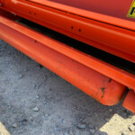Close-up of orange industrial equipment frame with bolts and a yellow warning label on a rocky dirt surface.