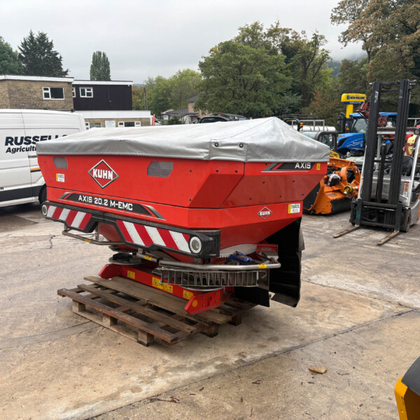 Red KUHN Axis 20.2 M-EMC agricultural machine covered with a tarp in a yard, with a white Russell Agriculture van and forklifts nearby.
