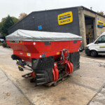 Red agricultural machine with a gray cover sits on pallets in a farm equipment yard outside a New Holland Service building with yellow and blue signage.