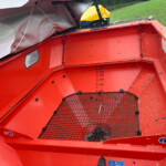 Red farm equipment hopper interior with a black grid floor and a vertical measurement scale on the side; a green field is visible outside.