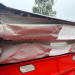 Close-up of a damaged red vehicle exterior with a torn gray cover, exposed wiring, and bent panels nearby.