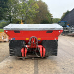 Front view of a large red farming implement with a gray cover in an outdoor equipment yard, with pallets and yellow attachments to the left and a blue tractor near a corrugated metal building to the right.