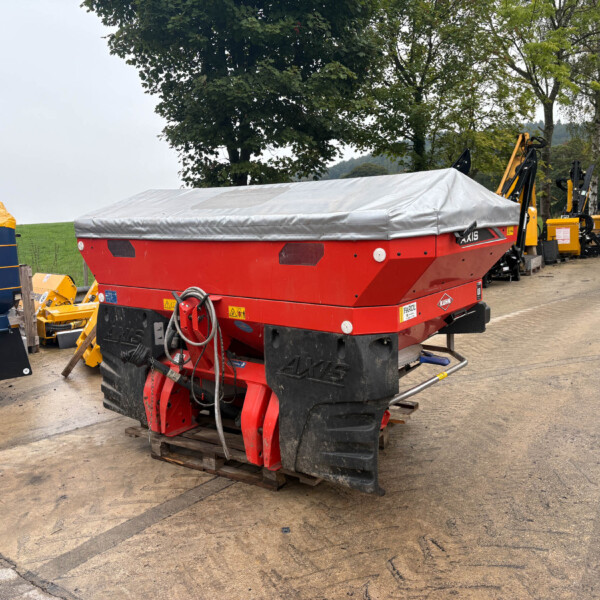 Red agricultural machine with a silver-covered tarp in a yard of yellow farming equipment in the background.