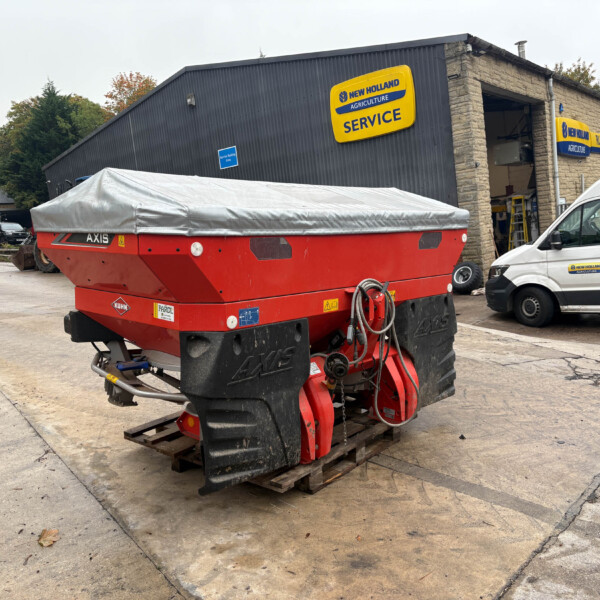 Red agricultural machine with a gray cover sits on pallets in a farm equipment yard outside a New Holland Service building with yellow and blue signage.