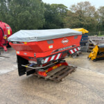Red KUHN Axis 20.2 M-EMC baler with a gray tarp cover, parked in a rural equipment yard beside yellow attachments and pallets behind it