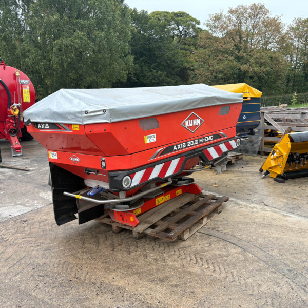 Red KUHN Axis 20.2 M-EMC baler with a gray tarp cover, parked in a rural equipment yard beside yellow attachments and pallets behind it