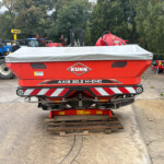 Red KUHN Axis 20.2 M-EMC agricultural trailer in the foreground, with blue New Holland tractors and various red farm equipment in a yard, and trees in the background, plus a wooden pallet on the ground.