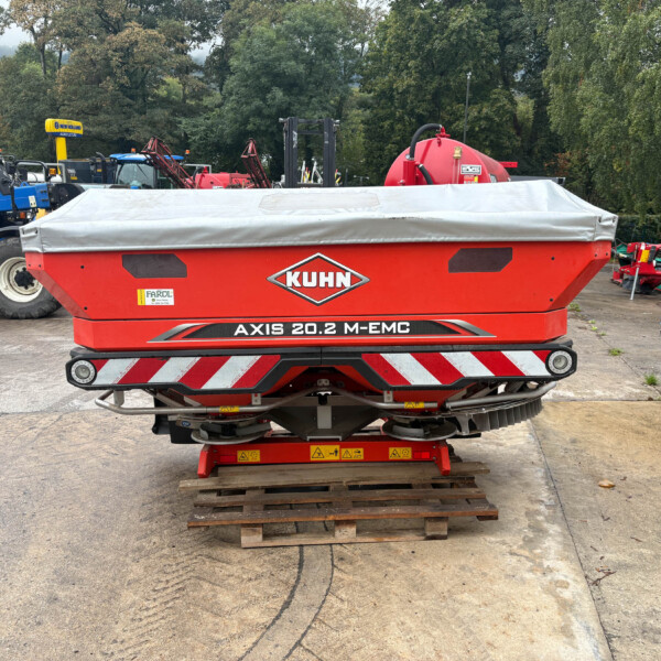 Red KUHN Axis 20.2 M-EMC agricultural trailer in the foreground, with blue New Holland tractors and various red farm equipment in a yard, and trees in the background, plus a wooden pallet on the ground.
