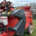 Red farm baler with hydraulic hoses and metal chute, parked on a grassy field with a fence and leafless trees in the background