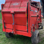 Red KUHN agricultural baler/wagon on a grassy field with hay sticking to its sides and a blue sky above