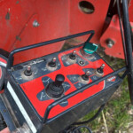 Red agricultural machine control panel with multiple toggles, levers, and a green dial; wiring visible nearby.