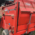 Red Kuhn Primor 3570 M baler with hay sticking to its surface, parked beside a trailer on a farm yard.