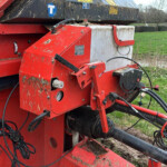 Close-up of red agricultural baler with hoses and wiring, dirt on metal, field in background.