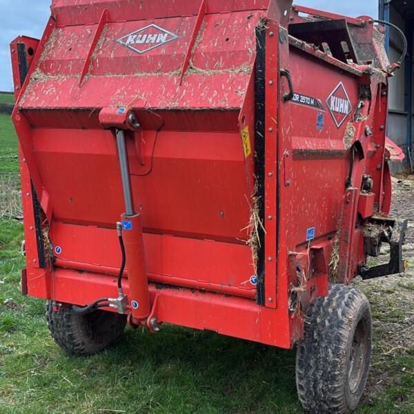 Red KUHN agricultural baler/wagon on a grassy field with hay sticking to its sides and a blue sky above