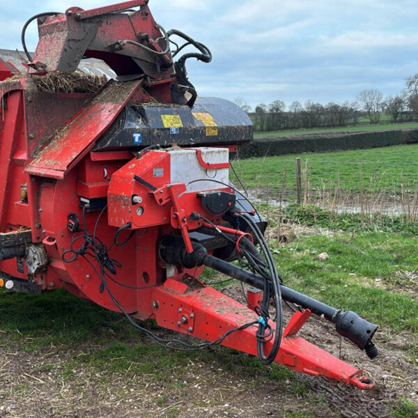 Red agricultural baler-like machine with hydraulic hoses sits on a grassy field, rural landscape in the background and overcast sky.