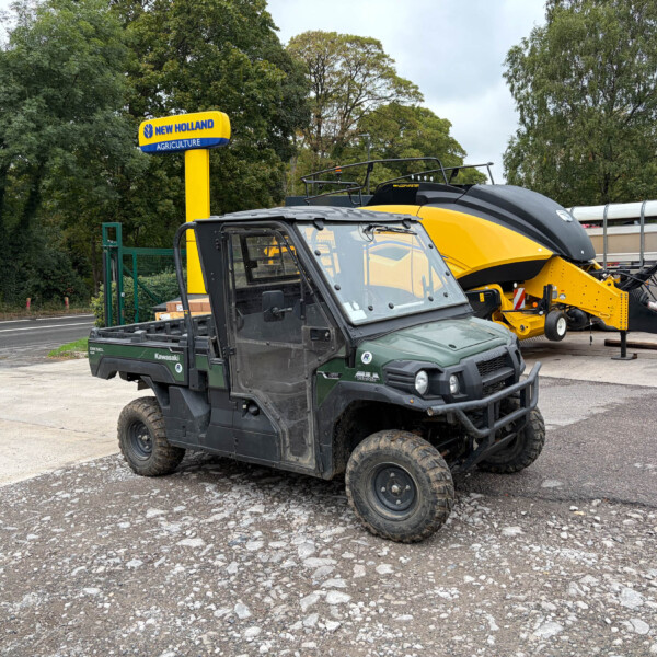 Yellow New Holland Agriculture sign beside a yellow agricultural machine in a parking lot with trees in the background, indicating an outdoor equipment display.