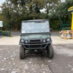 Front view of a gray off-road vehicle with oversized tires parked in a gravel lot, with construction gear and a yellow New Holland display in the background.