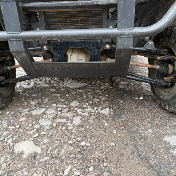 Underside of an off-road vehicle showing front differential, suspension, and large muddy tires, with a protective skid plate and steel bumper visible under a rocky surface.