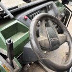 Close-up of the steering wheel and dusty dashboard of a green tractor, with red hazard button and various switches visible on the dash.