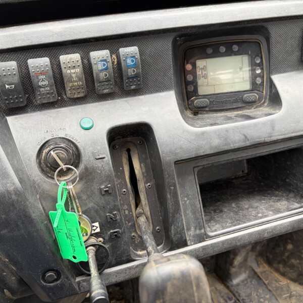 Cockpit of a dusty vehicle with multiple labelled rocker switches and an ignition key hanging on a ring. Green tags dangle from the keyring.