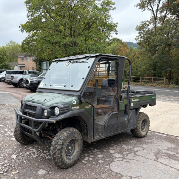 Green Kawasaki utility terrain vehicle parked in a car lot with trees and other vehicles in the background.