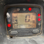 Kawasaki motorcycle digital gauge with speed readout at 0 mph, red warning lights on, and odometer reading visible; dusty surface around the display.