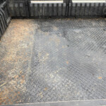 Truck bed with diamond-plate metal floor, dusty dirt, and scattered leaves along the edges and corners.