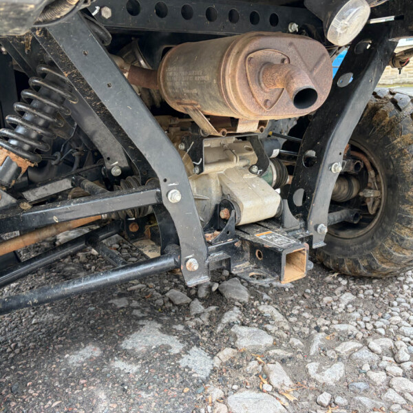 Rusty exhaust muffler and suspension under an off-road vehicle, with a dirty, rocky ground surface nearby.