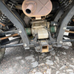 Underside of a dusty, rusted trailer showing coil springs, a rusty exhaust, and chassis beams around wheels.