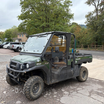 Front-left view of a Kawasaki Mule utility vehicle parked in a parking lot with trees in the background of a cloudy day.