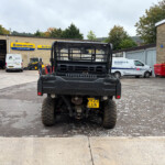 Rear view of a black Kawasaki utility truck in a service yard with a New Holland Agriculture sign and parked vans in the background.