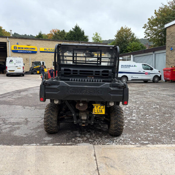 Rear view of a black Kawasaki utility truck in a service yard with a New Holland Agriculture sign and parked vans in the background.