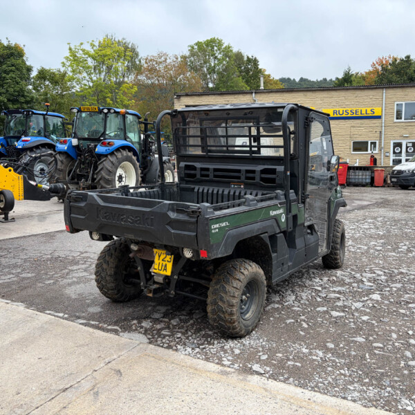 Black Kawasaki utility pickup with open cargo bed in a machinery yard; blue tractors and a beige building with a Russell’s sign in the background