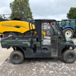 Bright yellow New Holland BigBaler 890 Plus baler parked in an outdoor equipment yard, with forklifts and a blue tractor nearby.
