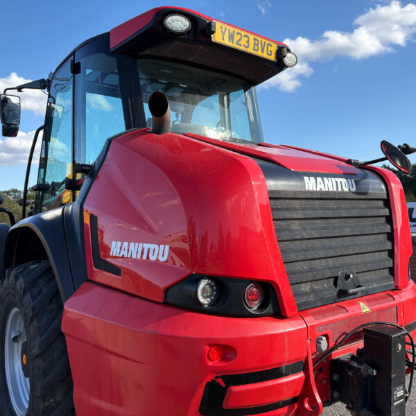 Close-up of a red Manitou tractor front with grill, headlights and a visible license plate (YW23 BVG) against a blue sky.