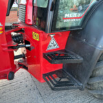 Close-up of red heavy machinery frame with a yellow safety guard and warning decals near a treaded wheel edge.