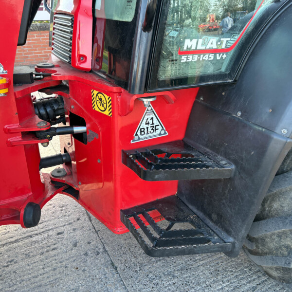 Close-up of red heavy machinery frame with a yellow safety guard and warning decals near a treaded wheel edge.