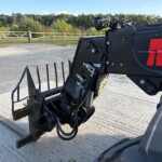 Close-up of a large black hydraulic construction attachment with hoses, on a paved surface, fence and trees in the background.